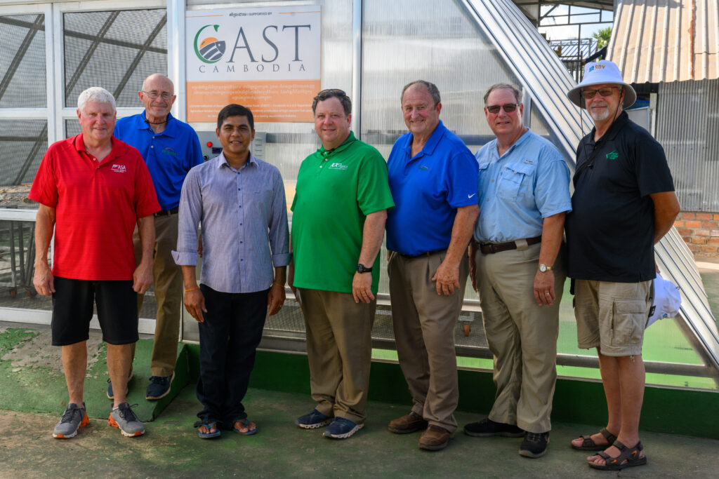 Six U.S. soybean farmers pose with a Cambodian fish processor in front of a solar dryer dome that dries fish.