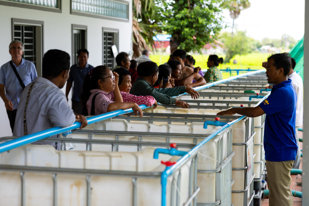 A man speaking to a group of people standing next to containers used for fish nurseries.