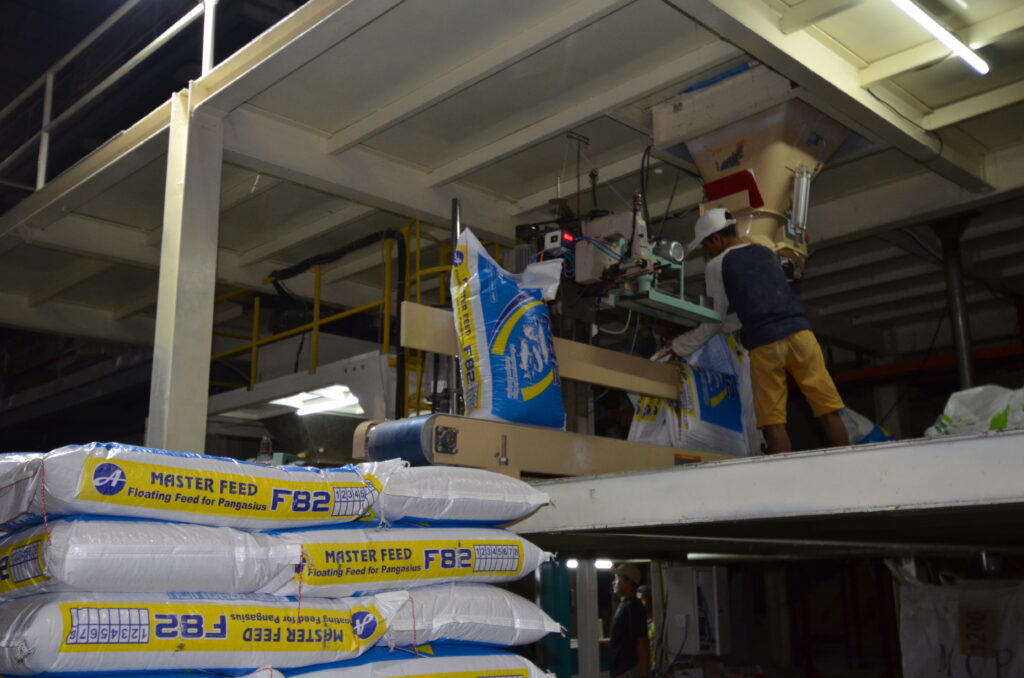 A man operating a piece of machinery at a feed mill with animal feed bags in the bottom left corner.