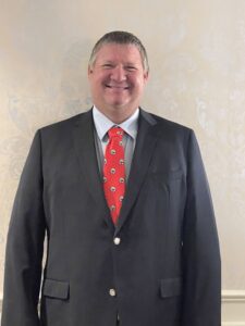A man smiling in a suit and tie stands in front of a wall.