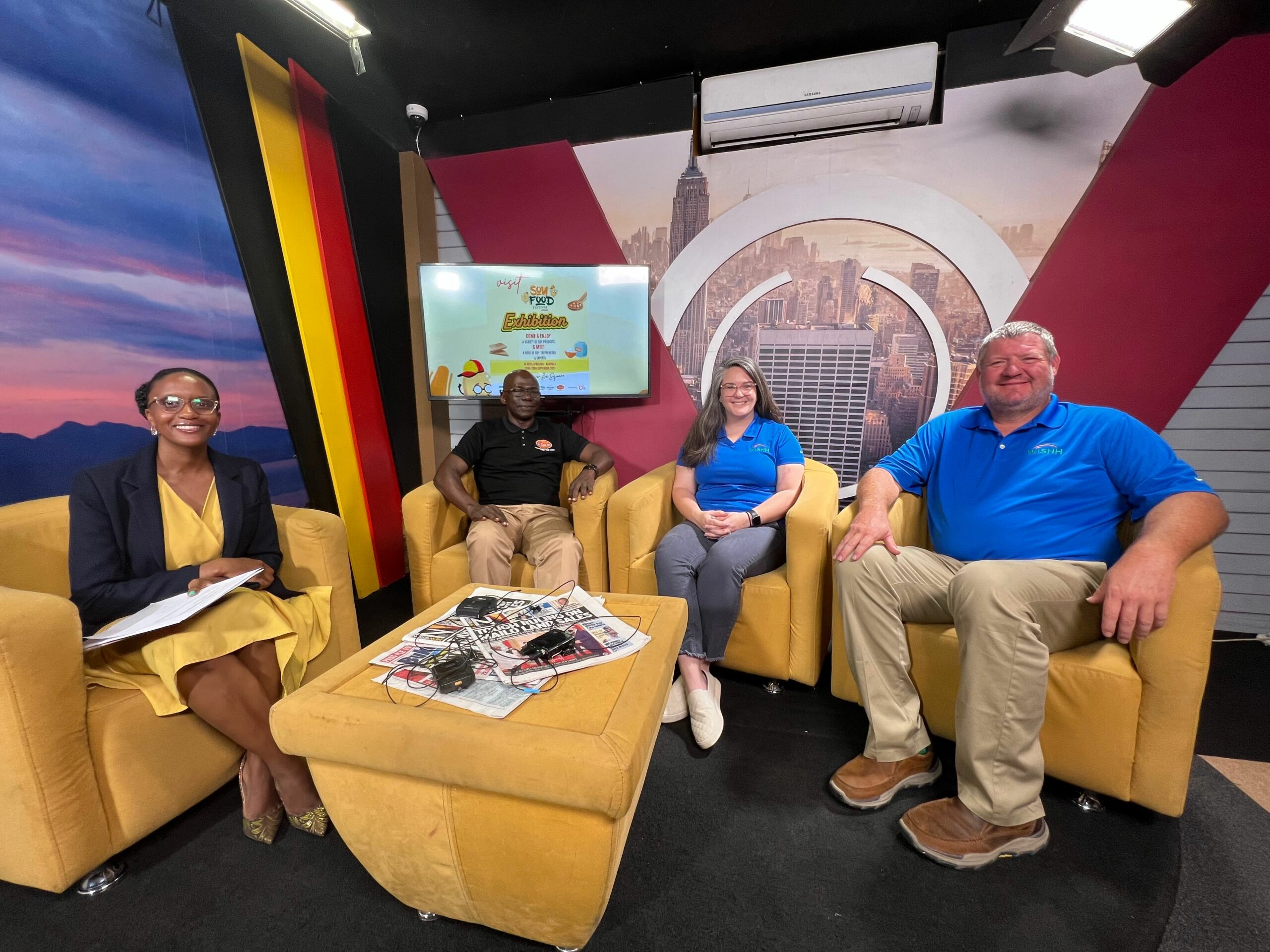 From right to left, Daniel Adams, Gena Perry, Charles Nsubuga, and a newscaster sit in seats around a coffee table on a news set.