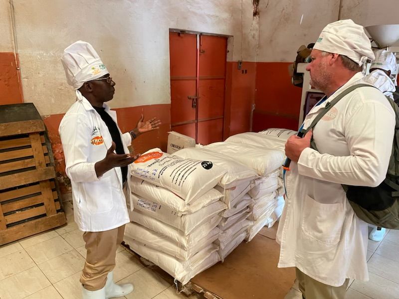 Two men wearing SESACO uniforms with chef hats stand speaking in front of bags filled with soy.