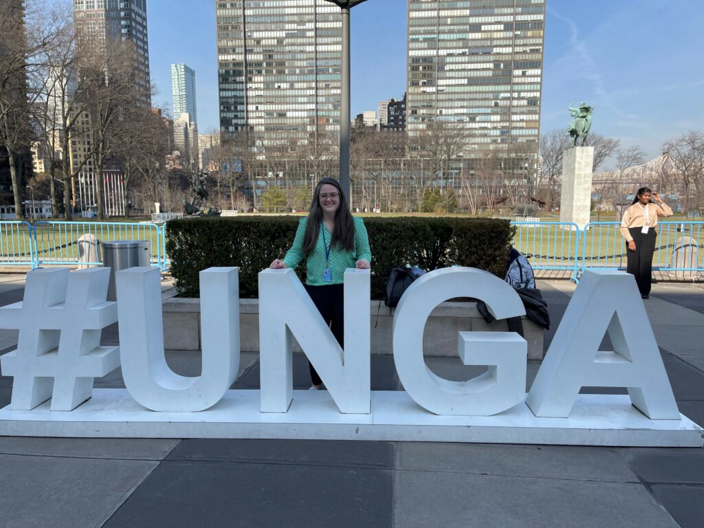 A woman standing outside behind a sign that reads as "#UNGA."
