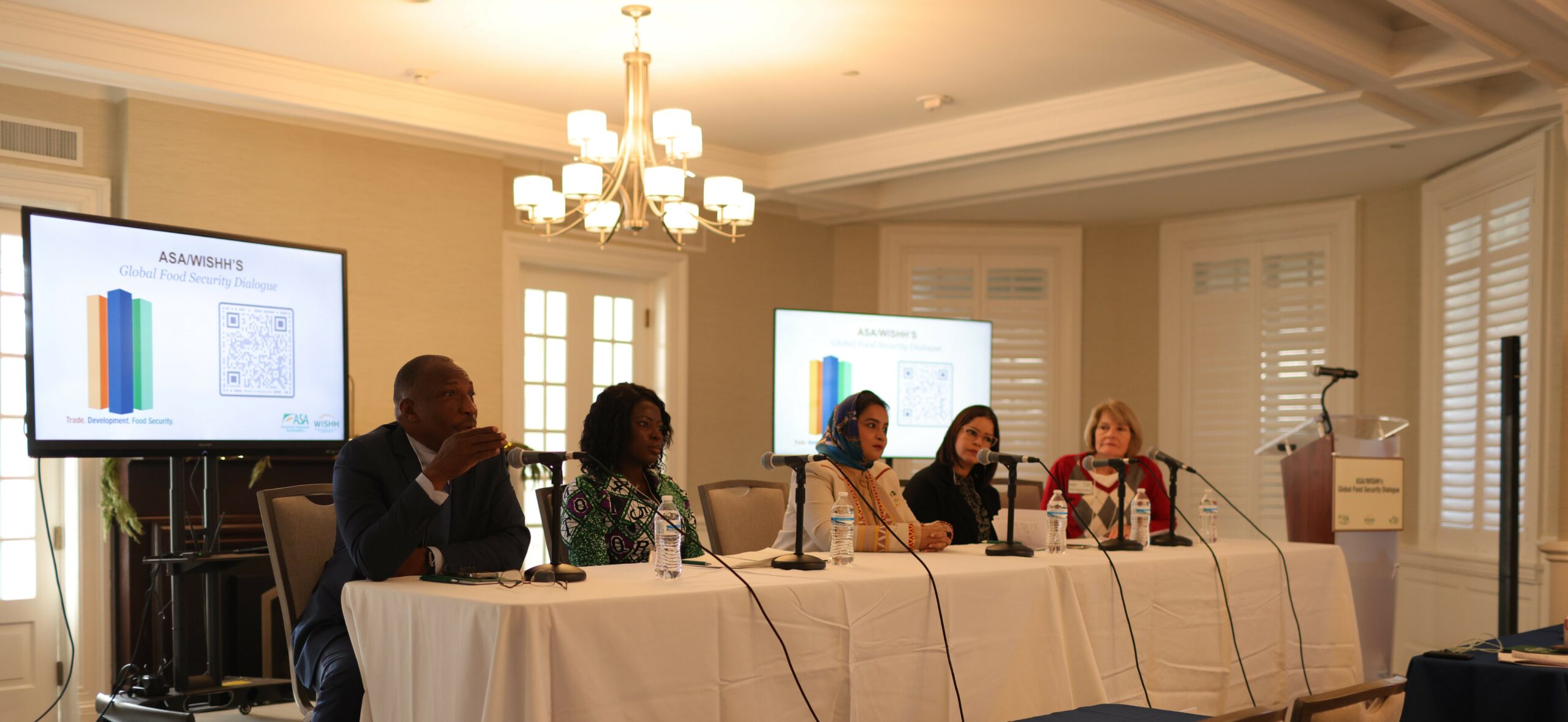 Five people sitting behind a long table with a white tablecloth and microphones in front of each of them.