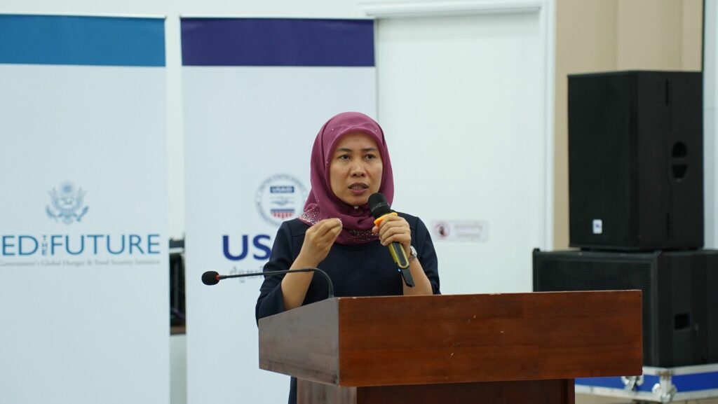 A Cambodian woman stands behind a podium with a microphone with banners in the background.