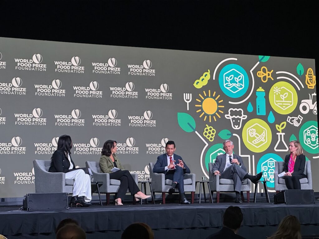 Five panelists sitting in grey chairs on a stage. The World Food Prize Foundation logo is in the background along with an agriculture graphic of different crops.