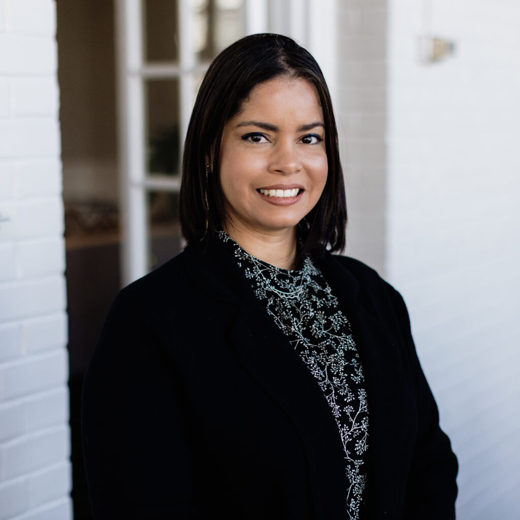 A woman, Elsy Alvarez, poses in front of a white gate in a business suit.