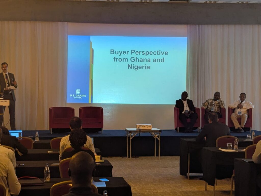 A man stands at a podium on the left side of a stage while three men sit in chairs on the right side of the stage. A screen projection on the back wall reads "Buyer Perspective from Ghana and Nigeria."
