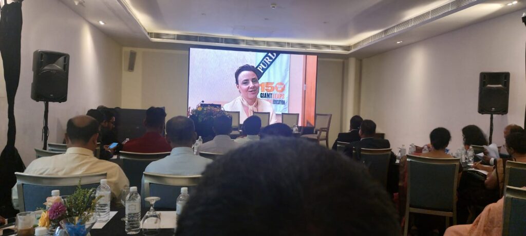 A video of a woman is shown projected against a wall in a room full of people sitting down and listening to the woman's virtual presentation.