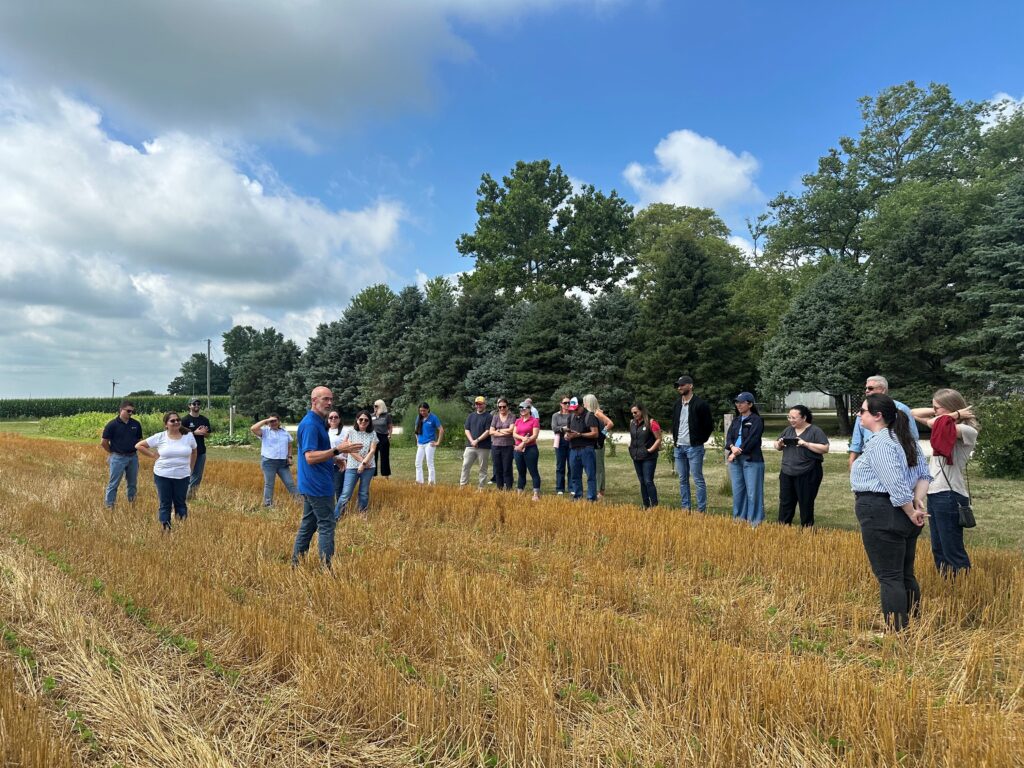 A farmer stands in a field while he speaks to a group of people.