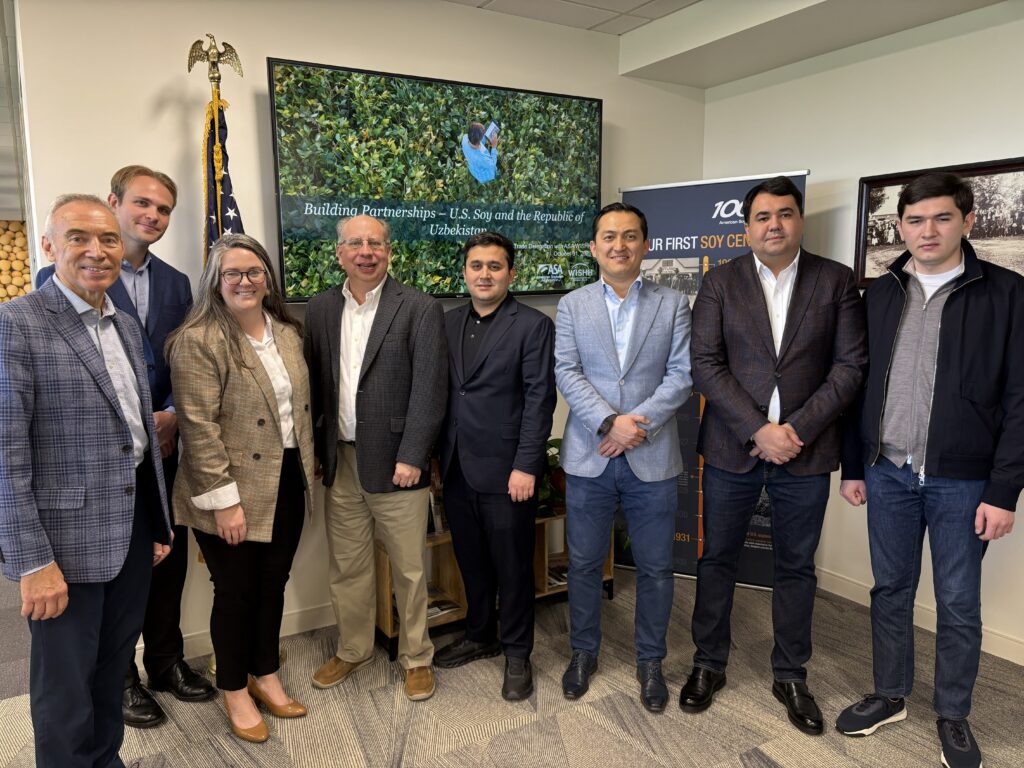 Eight people dressed formally stand side by side in front of a wall with a TV affixed to it.