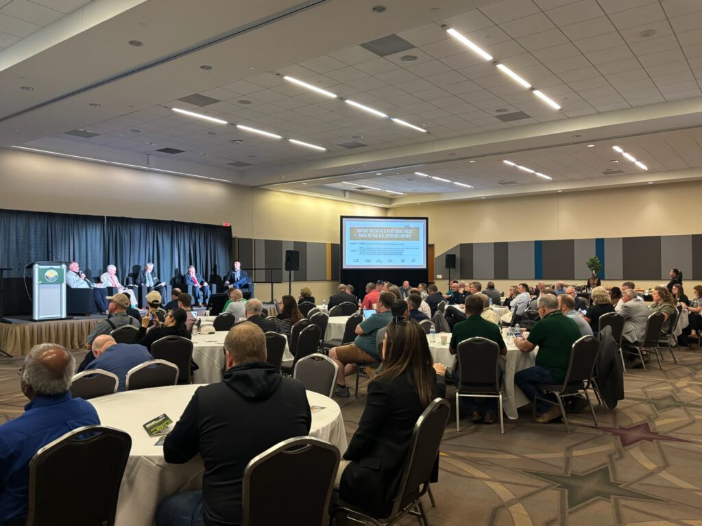 A room of people sitting at round tables while listening to panelists speaking while sitting in seating on top of a stage.