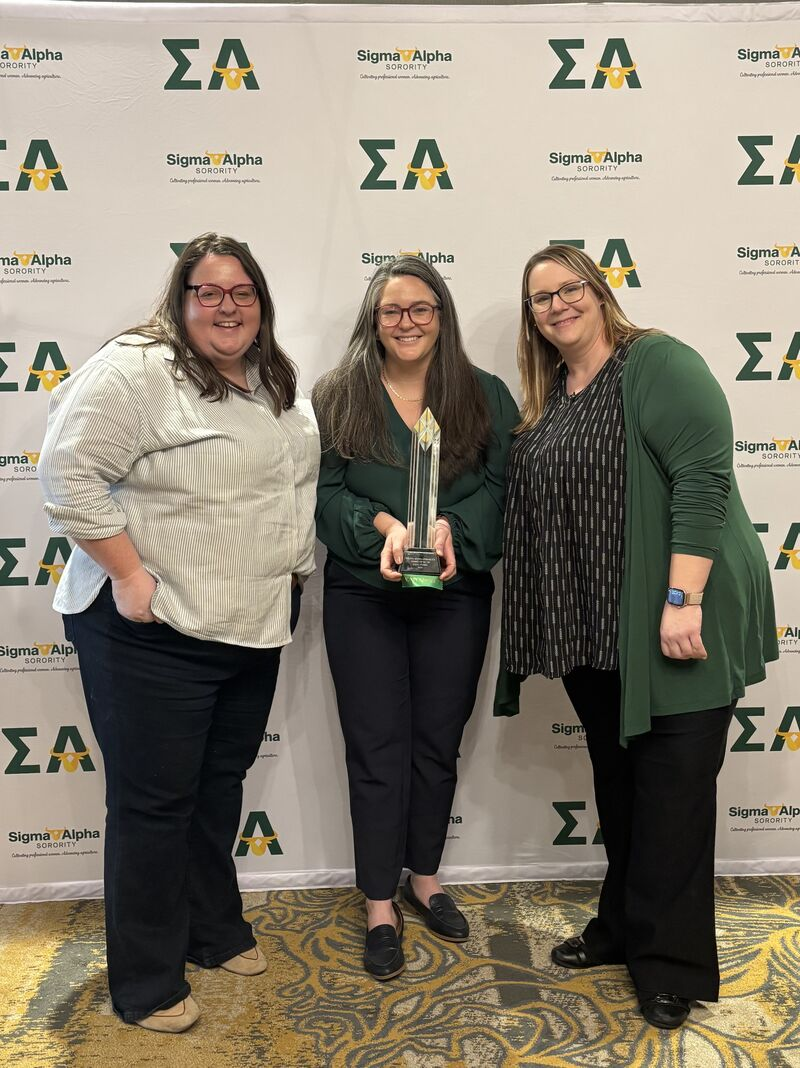 Three women post for a photo while the woman in the middle holds an award.