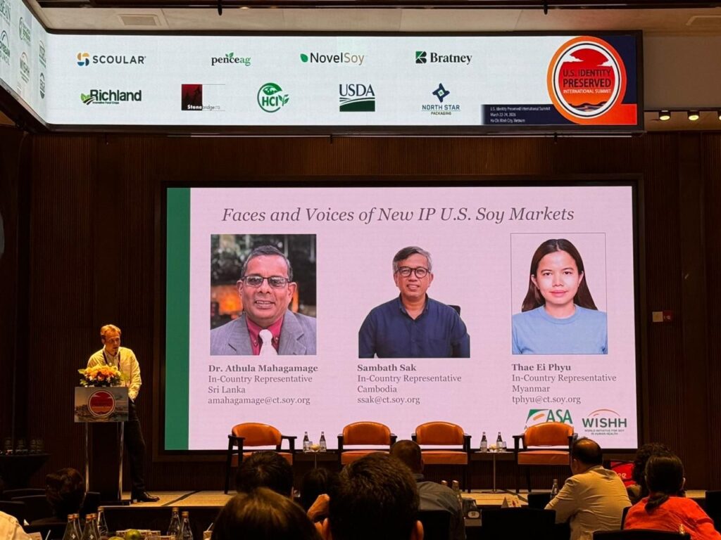 A man stands behind a podium to speak with a large projector screen in the background.