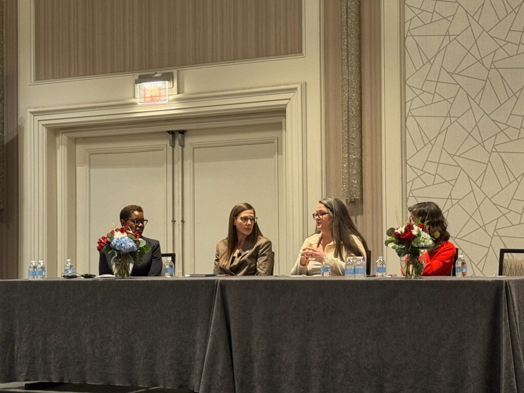Four women sit next to each other conversing behind a table with a gray tablecloth.